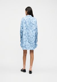 Γυναίκα που έχει γυρισμένη την πλάτη της, φοράει μια ανοιχτογαλάζια φόρεμα πουκάμισο με λουλουδένιο σχέδιο και μακριά μανίκια, ροδακινί κάλτσες και μαύρα μοκασίνια σε λευκό φόντο.