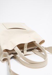 Bolso tote beige hecho de material suave con dos asas, un interior con cremallera y un bolsillo frontal plano. Diseño simple y funcional.