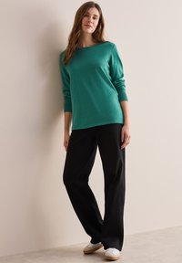 Türkise Langarmpullover mit weitem Ausschnitt, kombiniert mit schwarzen weit geschnittenen Hosen und weißen Sneakers vor einem hellen Hintergrund.