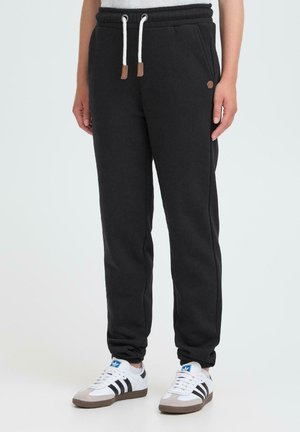 Person som har på seg svarte joggebukser med hvite snorer og hvite Adidas sneakers med svarte striper på en enkel bakgrunn.