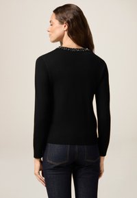 Zwarte gebreide trui met lange mouwen, voorzien van een halslijn met kraaldetail. Strakke pasvorm, aangevuld met donkerblauwe jeans.