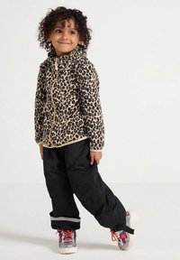 Ungdommelig barn med krøllete hår iført en leopardmønstret jakke med glidelås, svarte bukser og fargerike joggesko, stående med det ene benet krysset frem.