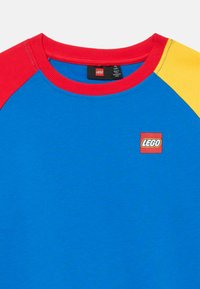 Μπλέ φούτερ με κόκκινες και κίτρινες ραγκλάν μανίκες, διαθέτοντας ένα μικρό κόκκινο λογότυπο LEGO στο στήθος. Φτιαγμένο από μείγμα βαμβακιού.