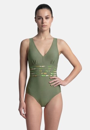 Femme aux cheveux foncés portant un maillot de bain une pièce vert avec un détail de taille rayé en maille, debout avec une main sur la hanche sur fond blanc.