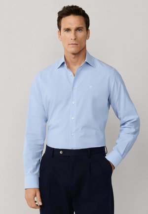 Man met blauwe ogen die een lichtblauw overhemd met knopen en donkerblauwe pantalon draagt, staand met één hand in de zak tegen een effen achtergrond.