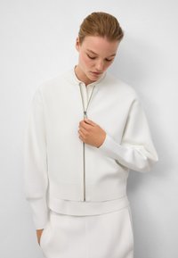 Veste zippée blanche avec col montant, fabriquée en tissu texturé. Présente des poignets et un ourlet côtelés. Le modèle tient la fermeture éclair dans une posture décontractée.