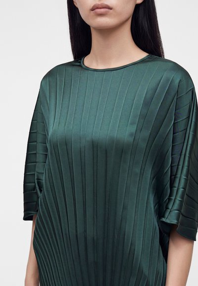 Femme portant un chemisier en satin vert foncé avec des plis verticaux et des manches larges arrivant aux coudes, visible des épaules à la taille.