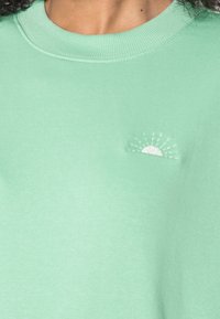 Sudadera verde menta con cuello acanalado y un pequeño diseño de sol bordado en blanco. La tela parece suave con una textura lisa.