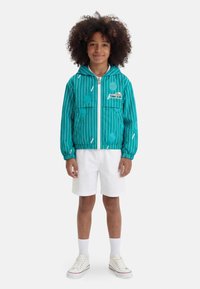 Παιδί με τζελ πράσινο μπουφάν με κουκούλα και σχέδια τένις, λευκά σορτς, κάλτσες και λευκά αθλητικά παπούτσια, που στέκεται μπροστά σε λευκό φόντο.