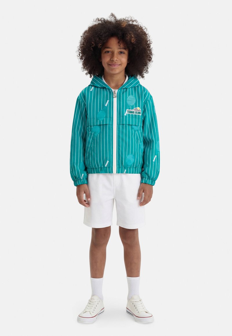 Παιδί με τζελ πράσινο μπουφάν με κουκούλα και σχέδια τένις, λευκά σορτς, κάλτσες και λευκά αθλητικά παπούτσια, που στέκεται μπροστά σε λευκό φόντο.