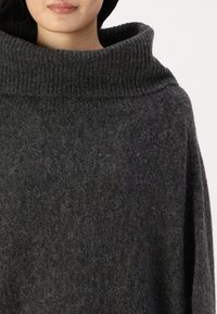 Pull en maille de couleur gris foncé, oversized, avec un col montant épais à côtes et un tissu doux et texturé. Il présente des manches larges et une coupe décontractée.