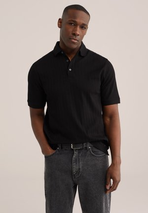 Mladý muž stojaci s rukami v vreckách, oblečený v čiernej polokošeli s krátkym rukávom a vyblednutých čiernych rifle na jednoduchom pozadí.