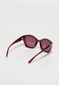 Καρμινί γυαλιά ηλίου με γωνιώδεις, oversized σκελετούς και φακούς με γραδιέντο. Διαθέτουν χρυσή λεπτομέρεια στη βάση, με γυαλιστερή φινίρισμα.