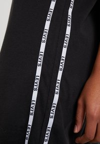 Vêtement noir Levi's avec deux cordons verticaux blancs ornés du logo Levi's répété, à côté de la main droite détendue d'une personne.