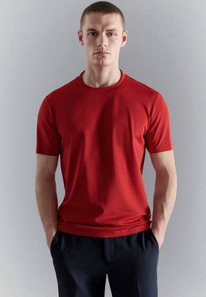 Giovane uomo con capelli corti che indossa una maglietta rossa semplice a maniche corte e pantaloni scuri, in piedi davanti a uno sfondo grigio chiaro.