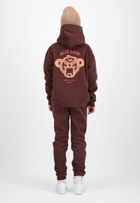 Persoon draagt een bruine hoodie en broek met een abstracte aapdesign en tekst op de achterkant, beige gebreide muts, witte sneakers, staand op een witte achtergrond.