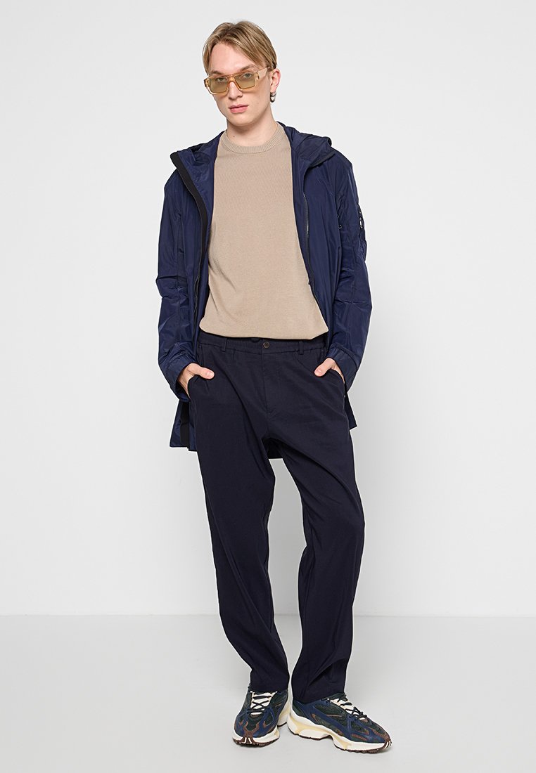 Granatowa wodoodporna kurtka, beżowy sweter i ciemne spodnie. Szaro-niebieskie trampki. Przezroczyste okulary. Stoi na neutralnym tle.