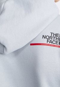 Tejido de color gris claro con textura plegada y el logo de "The North Face" en negro por encima de una línea roja con texto pequeño en blanco.