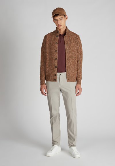 Jonge man in een bruine gebreide cardigan, bordeauxkleurig poloshirt, beige broek, witte sneakers en een bruine pet, staand tegen een effen achtergrond.