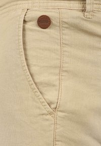 Beige-huppu-kangasta tehty housu, jossa on sivutasku, teksturoitu kangas ja ruskea nahka-akkentu, jossa on kuvattu brändin nimi.