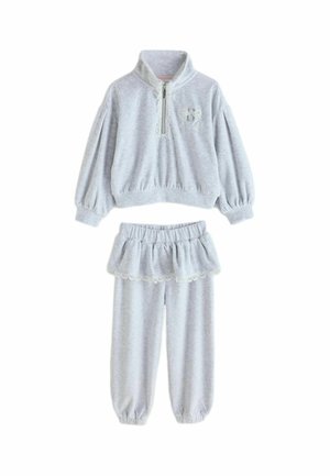 Young girl wearing light gray sweatshirt and pants set with lace trim and white shoes with pink bows standing on gray floor.