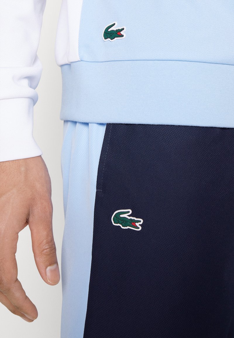 Sudadera azul claro con un panel blanco, combinada con joggers marinos. Presenta el logo de Lacoste en ambas prendas. Tejido suave, puños y cinturilla acanalados.
