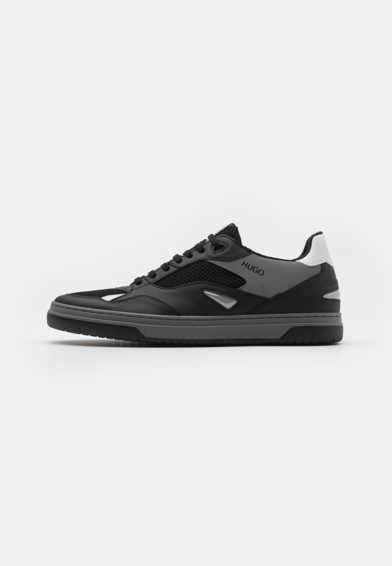 Baskets de sport noires avec un mélange de mailles et de cuir synthétique, présentant un profil bas, un bout rond et des accents gris. Lacets et logo visibles.