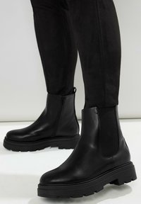 Svarta läderankelskor med en kraftig gummisula och elastiska sidopaneler. Slät yta med rundad tådesign. Dragkedjedetalj på ena sidan.