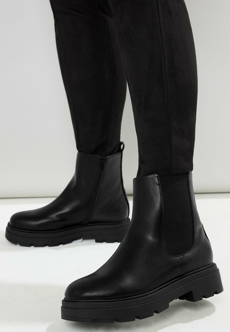 Svarta läderankelskor med en kraftig gummisula och elastiska sidopaneler. Slät yta med rundad tådesign. Dragkedjedetalj på ena sidan.