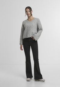 Šedý svetr s kabelovým vzorem a výstřihem do V, kombinovaný s rozšířenými černými džínami a světlými teniskami. Textura je hrubá s viditelnými vzory švů.