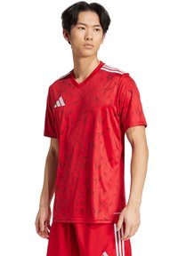 Maglia da calcio rossa con un motivo geometrico, design a V, maniche corte e strisce bianche sulle spalle. Realizzata in tessuto leggero.