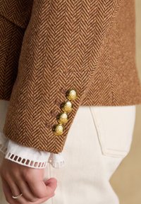 Manche de blazer marron en chevrons avec quatre boutons décoratifs dorés sur un poignet orné de dentelle blanche, à côté d'une poche de pantalon beige.