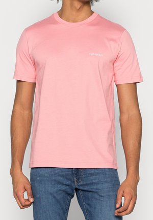 Mand iført en enkel lyserød kortærmet t-shirt med lille hvid Calvin Klein-logo på brystet, kombineret med blå jeans.