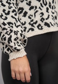 Leopardmønstret genser med en kremfarget bakgrunn, svarte mønstre og ribbede mansjetter. Nærbilde av ermet og hånden mot svarte leggings.