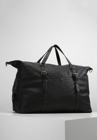 Bolso de viaje de cuero negro con acabado texturizado, doble asa en la parte superior, correas ajustables y bolsillo frontal. Los acentos incluyen herrajes en bronce.