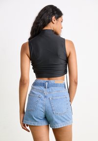 Zwarte mouwloze crop top met een hoge hals en geplooide details, gecombineerd met lichtblauwe hoog-getailleerde denim shorts met twee achterzakken.