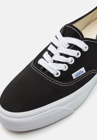 Baskets Vans noires avec lacets blancs, coutures blanches et semelle en caoutchouc blanche, affichant le logo en tissu Vans sur le côté.