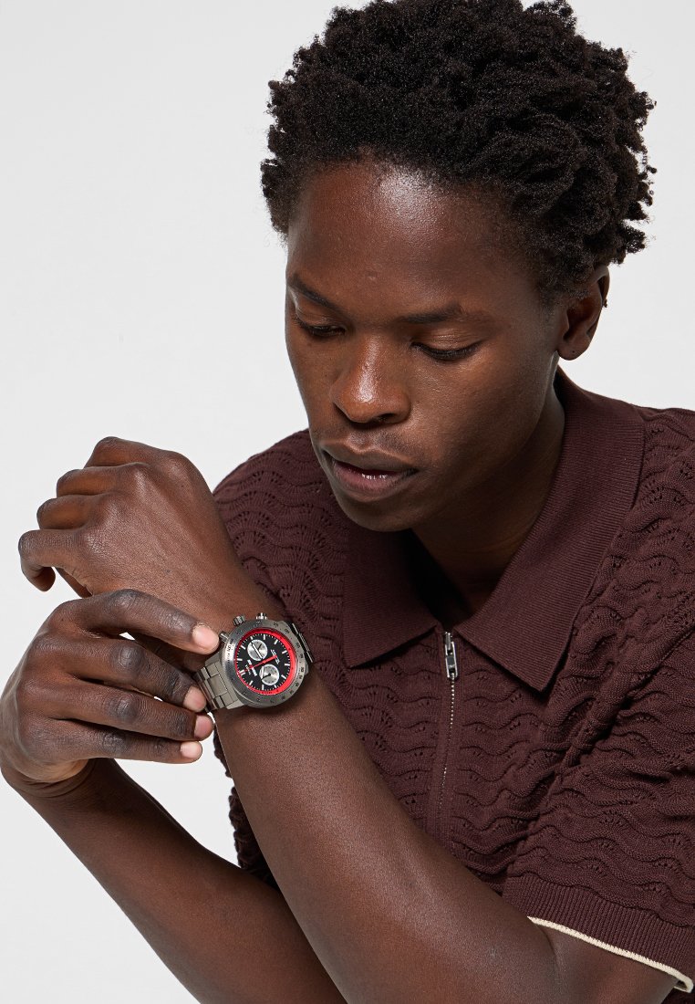 Hombre ajustando un reloj de pulsera metálico con esfera negra y roja, vistiendo una camisa marrón con estampado de manga corta y cuello con cremallera.