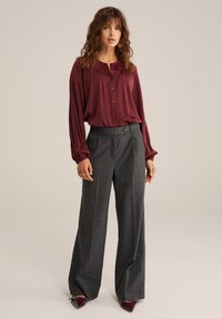 Donna che indossa una camicia button-down a maniche lunghe color bordeaux, pantaloni larghi grigi e scarpe a punta bordeaux, in piedi su uno sfondo semplice.