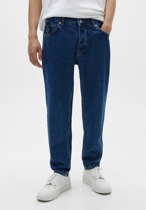 Osoba ubrana w luźne, ciemnoniebieskie jeansy, białe sneakersy i białą koszulę, stojąca z rękami w kieszeniach.