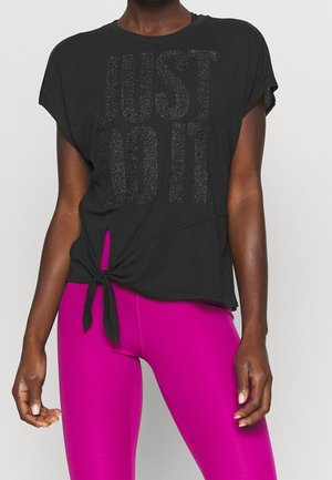 Persoon draagt een zwart vastgeknoopt shirt met de tekst "Just Do It" en felroze leggings, staand tegen een effen witte achtergrond.