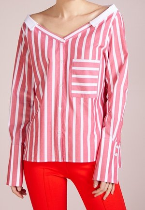 Femme portant un chemisier à rayures verticales roses et blanches avec une poche carrée rayée et un pantalon rouge vif.