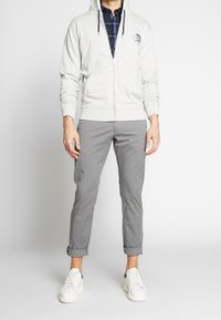 Hellgraue Reißverschlussjacke mit Logo auf der Brust, kombiniert mit grauen Hosen mit umgeschlagenen Säumen und weißen Sneakers. Lässiges Outfit mit übereinanderliegenden Texturen.