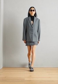 Grijze blazer over een zwarte turtleneck, gecombineerd met een gemusterde minirok en chunky zwarte schoenen. Model draagt donkere zonnebril en staat op een houten vloer.