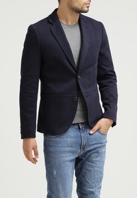 Navy blazer made from fabric with a smooth texture, featuring a single button closure, notch lapels, and a slim fit design. Worn over a gray shirt.