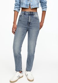 Personne portant un jean bleu délavé taille haute, une veste courte en denim et des baskets blanches sur un fond blanc.