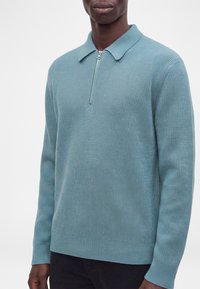 Homme portant un pull en maille côtelée bleu clair avec une courte fermeture éclair et un col, associé à un pantalon foncé sur un fond uni.