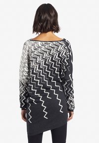Kobieta z krótkimi ciemnymi włosami nosi długi sweter w czarno-białą zygzakowatą wzór, stojąc tyłem na tle gładkiej białej ściany.