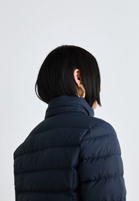 Navy puffer jacket with a high collar, featuring quilted texture and a smooth finish, viewed from the back. Gold hoop earring visible.