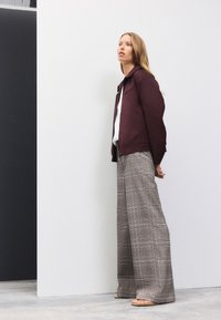 Giacca bordeaux con collo a risvolto, abbinata a pantaloni a gamba larga a quadri in tonalità di grigio, con sottili accenti rossi e una texture liscia.